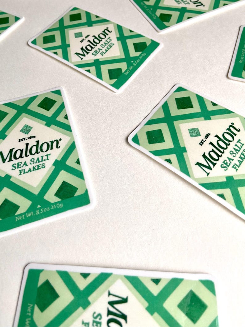 Może przedstawiać: Kilka opakowań Maldon Sea Salt Flakes rozrzuconych na białej powierzchni. Każde opakowanie ma zielono-biały geometryczny wz&oacute;r, nazwę marki i szczeg&oacute;ły produktu. Waga netto wynosi 240g.