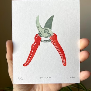 May include: A print of red and green pruning shears on white paper. The word "pruners" is written in black script below the shears. The print is signed and numbered 2/OE.