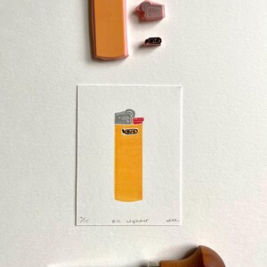 Op de afbeelding: Een print van een gele BIC-aansteker met de tekst "BIC Lighter" en het nummer "4/15". De afbeelding bevat ook een snijgereedschap en diverse rubberen stempels.