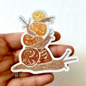 May include: A sticker of a stack of four snails, with the largest snail at the bottom and the smallest snail on top. The snails are drawn in shades of brown and yellow.