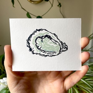 May include: A small, rectangular art print featuring a stylized oyster illustration. The oyster is depicted in shades of green and black against a white background. The print is held in a hand, with greenery visible in the background.
