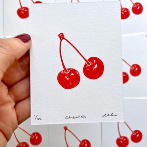 May include: A red and white print of two cherries with stems on a white background. The print is signed "Cherries" and numbered "1/06".