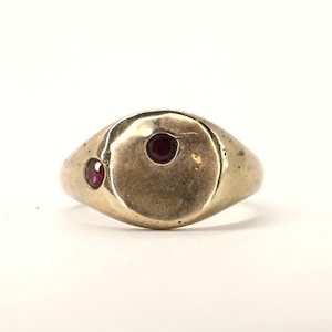 May include: A gold-toned metal signet ring featuring a smooth, rounded face with a textured, organic finish. The ring is set with two small, circular, deep red gemstones embedded asymmetrically into the surface of the metal band.