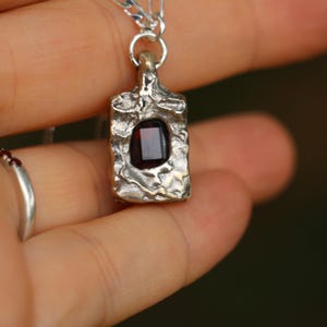 May include: Silver pendant with a rectangular, textured frame and a dark red, square gemstone. The pendant hangs from a silver chain. The design has a rustic, handcrafted appearance.