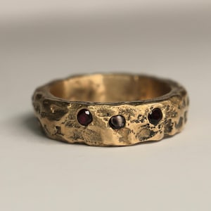 May include: A gold ring with a textured surface and three small, round, red gemstones. The ring has a rustic, uneven appearance. The gemstones are evenly spaced around the ring.