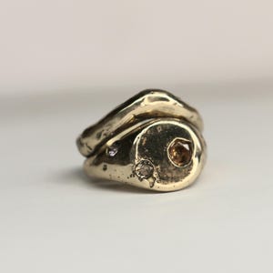May include: A gold-toned ring with a unique, organic shape. The ring features three small, sparkling gemstones: one clear, one brown, and one purple. The design is abstract and modern, with a textured surface.