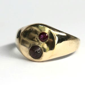 May include: A gold ring featuring two inset gemstones. One is a deep red, the other a darker, purplish-red. The ring has a smooth, rounded design. The gemstones are round and set into the gold. A piece of jewellery.