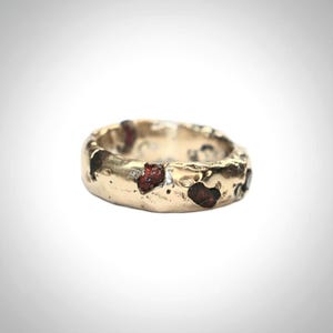 May include: A gold-toned ring with a textured, uneven surface featuring embedded dark red, irregular stone fragments. The band has a thick, organic shape with a metallic finish and visible indentations throughout the exterior circumference.