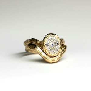 May include: A gold ring featuring a central oval-cut clear gemstone set in a textured, organic-shaped band. The metal has a warm, yellow-gold tone with a hammered, uneven surface finish that contours around the base of the oval stone setting.
