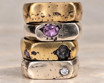 May include: A stack of four wide metal rings with a textured, rustic finish. The top ring is gold-toned, the second is silver-toned with a purple oval stone, the third is gold-toned with dark and purple accents, and the bottom is silver-toned with a clear stone.