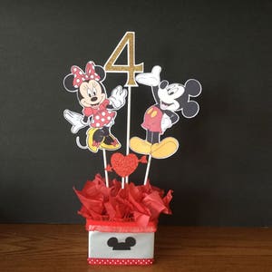 May include: A Mickey Mouse and Minnie Mouse themed centerpiece for a 4th birthday party. The centerpiece features a gold number 4, Mickey Mouse and Minnie Mouse characters, red hearts, and red tissue paper in a white box with a black Mickey Mouse head.