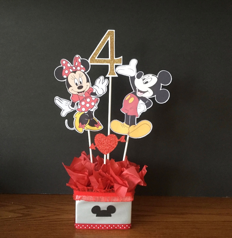 May include: A Mickey Mouse and Minnie Mouse themed centerpiece for a 4th birthday party. The centerpiece features a gold number 4, Mickey Mouse and Minnie Mouse characters, red hearts, and red tissue paper in a white box with a black Mickey Mouse head.