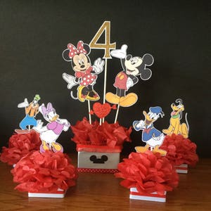 May include: Mickey Mouse and Minnie Mouse themed centerpiece with red tissue paper balls, a red heart, and a gold glitter number 4. The centerpiece is surrounded by four smaller centerpieces featuring Donald Duck, Goofy, Pluto, and Daisy Duck.