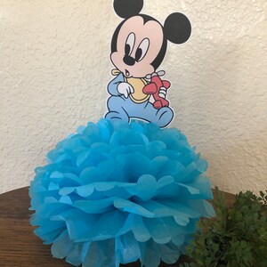 May include: Blue tissue paper pom pom with a baby Mickey Mouse character holding a red airplane on top. The character is wearing a blue onesie and a yellow bib.