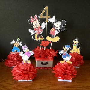 May include: A Mickey Mouse themed centerpiece for a 4th birthday party. The centerpiece features Mickey Mouse, Minnie Mouse, Donald Duck, Goofy, and Pluto characters. The characters are standing on red tissue paper pom-poms. The centerpiece also includes a red and white polka dot box with a Mickey Mouse head on it.