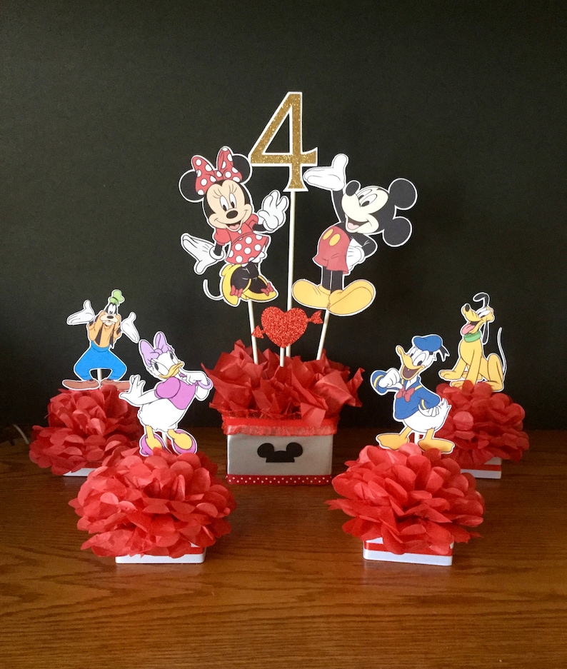 May include: A Mickey Mouse themed centerpiece for a 4th birthday party. The centerpiece features Mickey Mouse, Minnie Mouse, Donald Duck, Goofy, and Pluto characters. The characters are standing on red tissue paper pom-poms. The centerpiece also includes a red and white polka dot box with a Mickey Mouse head on it.
