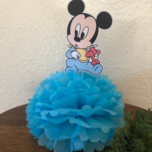 May include: Blue and white paper pom-pom with a baby Mickey Mouse character holding a red airplane on top. The character is wearing a blue onesie and a yellow bib.