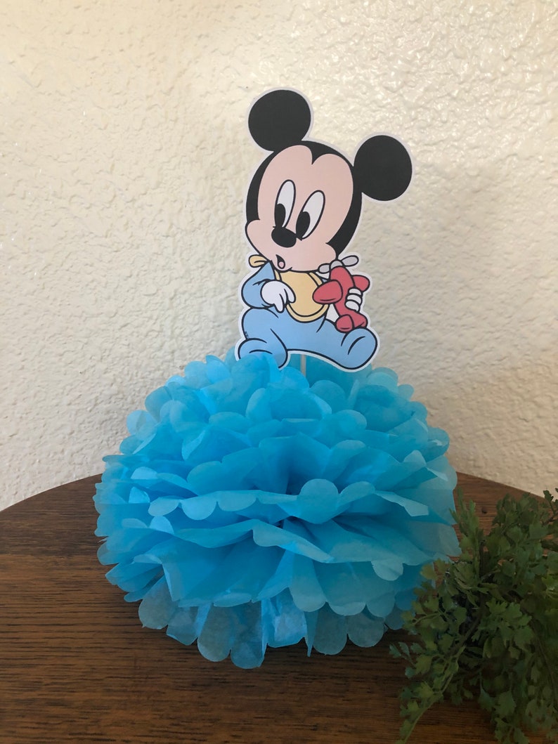May include: Blue and white paper pom-pom with a baby Mickey Mouse character holding a red airplane on top. The character is wearing a blue onesie and a yellow bib.