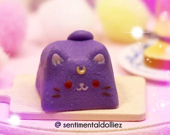 May include: A purple cat-shaped keycap with a gold crescent moon symbol on its forehead, pink cheeks, and whiskers, resting on a miniature wooden cutting board. The text @sentimentaldolliez is printed on the board surface.