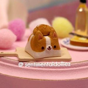 May include: A miniature pink table with a decorative corgi-shaped cake on a wooden cutting board. The table also features a small bottle, a slice of toast, and colorful pom-poms. The text "@sentimentaldolliez" is visible on the table.