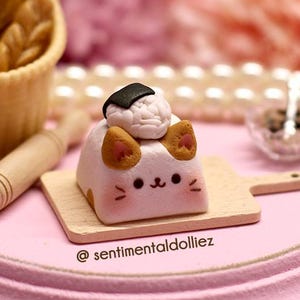 May include: A miniature scene with a pink wooden base and a white cat-shaped tofu with a seaweed-wrapped rice ball on top. The tofu is on a small wooden cutting board. There is a rolling pin, a small carton of milk, and a basket of bread in the background.