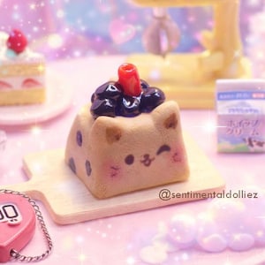May include: A miniature scene featuring a cat-shaped cake topped with dark berries and a red cherry, a slice of strawberry cake, and a yellow mixer. A pink heart-shaped counter and a small bowl of cookies are also present. The scene is set on a pink surface.