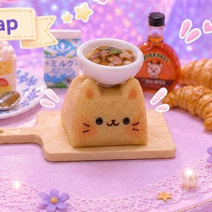 Puede incluir: Un caprichoso juego de teclas artesanales con un cuenco de pho con forma de gato, pastel y cruasanes sobre un soporte decorativo. El juego de teclas está rodeado de flores moradas y el texto "Kawaii CAT PHO".