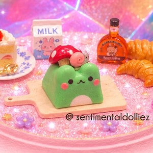 May include: A pink cake stand displays miniature desserts and pastries. A green frog-shaped cake with a mushroom, a slice of cake with a strawberry, a bottle of liquor, croissants, and a carton of milk are arranged on the stand. The text "@sentimentaldolliez" is visible.