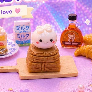 May include: A whimsical artisan keycap display featuring miniature food items like a cake slice, croissants, and a maple syrup bottle. The keycap has a cute character design. The scene is set on a purple platform with floral accents and the text "Kawaii Artisan Keycap".