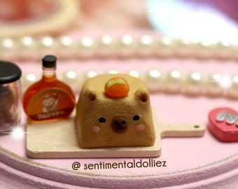 May include: A miniature wooden cutting board with a small brown bear-shaped cake, a bottle of maple syrup, and a jar of brown sugar. The cutting board is on a pink pedestal with a pearl necklace around it. The text "@sentimentaldollliez" is on the cutting board.