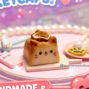 May include: A whimsical image showcasing handmade and kawaii keycaps. The keycaps are designed as cute bread shapes, including a cat-shaped bread with a smiling face. The image also features a pink platform and the text "Cute Bread Keycaps!"