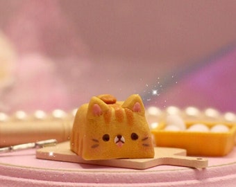 May include: A miniature, orange cat-shaped bread loaf with brown stripes and a cute face, resting on a small wooden cutting board. The scene is set on a pink, tiered stand with pearls and a rolling pin, suggesting a bakery theme.