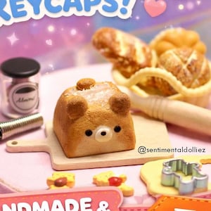 May include: A collection of handmade and kawaii bread-themed keycaps. The image features a bear-shaped bread keycap, miniature bread rolls, a rolling pin, and a small whisk. The text "Cute Bread Keycaps!" and "Handmade & Kawaii!" are displayed.