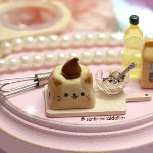 Puede incluir: Una escena de cocina en miniatura con un soporte para pasteles rosa, una pequeña tabla de cortar de madera, un batidor, un tazón de chispas marrones, una botella de aceite, una caja de harina y un lindo pastel con forma de gato con glaseado de chocolate.