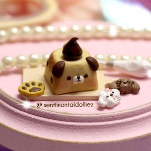 Puede incluir: Un pastel miniatura con forma de perro con glaseado de chocolate y una huella de pata en una pequeña tabla de madera. El pastel está en un soporte de pastel rosa con un collar de perlas y una manga pastelera.