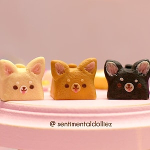 K&ouml;nnte beinhalten: Drei kleine, handbemalte Keramik-Tastenkappen in Form von Hunden. Die Tastenkappen sind in den Farben Beige, Braun und Schwarz gehalten. Die Tastenkappen liegen auf einer rosa Oberfl&auml;che. Der Text "@sentimentaldolliez" ist unter den Tastenkappen sichtbar.