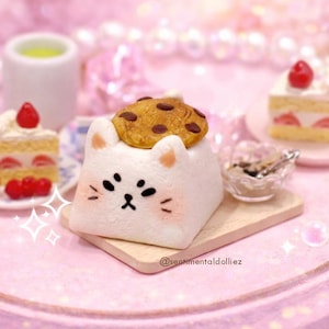 Puede incluir: Una escena de postres en miniatura caprichosa con un pastel en forma de gato cubierto con una galleta con chispas de chocolate. Otros dulces incluyen rebanadas de pastel de fresa y palitos de pan en miniatura, todo dispuesto en una bandeja rosa.