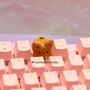 K&ouml;nnte beinhalten: Eine braune und goldene, hundeartige Tastenkappe sitzt auf einer rosa Tastatur mit wei&szlig;en Beschriftungen. Die Tastenkappe befindet sich auf der Taste "H". Die Tastatur hat den Text "@sentimentaldolliez" auf der Taste "G" aufgedruckt.