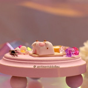 May include: A miniature pink and white cake with brown details, sitting on a pink wooden base with a gold trim. The base has three legs and the text "@sentimentaldolliez" is written on the front. There are also small pink and purple glass gems on the base.