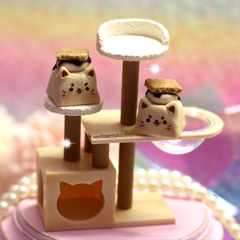 K&ouml;nnte beinhalten: Ein skurriles Miniatur-Katzenbaum mit zwei katzenf&ouml;rmigen Leckereien, die mit S'mores belegt sind. Der Katzenbaum ist beige mit einer wei&szlig;en Sitzfl&auml;che und einem katzenf&ouml;rmigen Ausschnitt. Die Leckereien haben ein s&uuml;&szlig;es Gesicht.