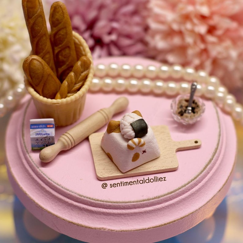 May include: A miniature scene featuring a pink table, a small wooden chopping board, a white cat figurine with a sushi roll on its head, a rolling pin, a small box of crackers, a basket of bread, and a small bowl of brown sugar.