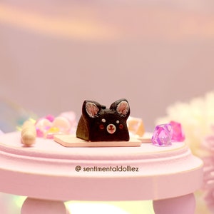 K&ouml;nnte beinhalten: Ein kleiner schwarz-wei&szlig;er Kuchen in Form eines Hundes vor einem rosa Hintergrund. Der Kuchen steht auf einem kleinen rosa Tisch mit einem wei&szlig;en Sockel.