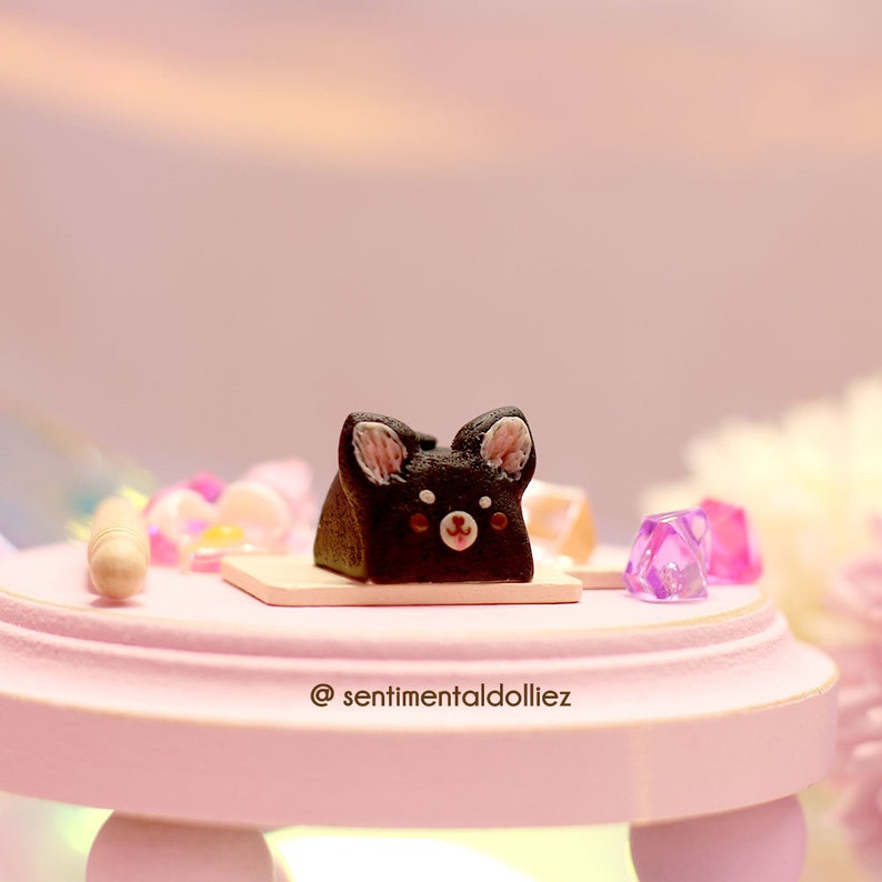 K&ouml;nnte beinhalten: Ein kleiner schwarz-wei&szlig;er Kuchen in Form eines Hundes vor einem rosa Hintergrund. Der Kuchen steht auf einem kleinen rosa Tisch mit einem wei&szlig;en Sockel.