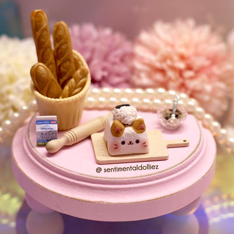 May include: A miniature cat-shaped cake with white and brown icing, sitting on a wooden chopping board. The cake is on a pink cake stand with a rolling pin and a small basket of bread rolls.