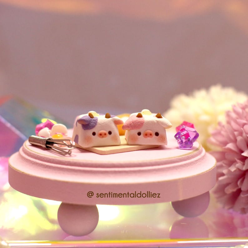 May include: Two miniature pink and white cow-shaped cakes with purple spots on a pink cake stand with a gold trim. The stand has three legs and is on a reflective surface. The text "@sentimentaldolliez" is on the stand.