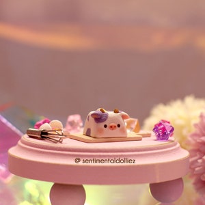 May include: A miniature purple and white cow figurine on a pink cake stand with a small whisk and a pink and purple crystal. The stand has the text "@sentimentaldolliez" on the front.
