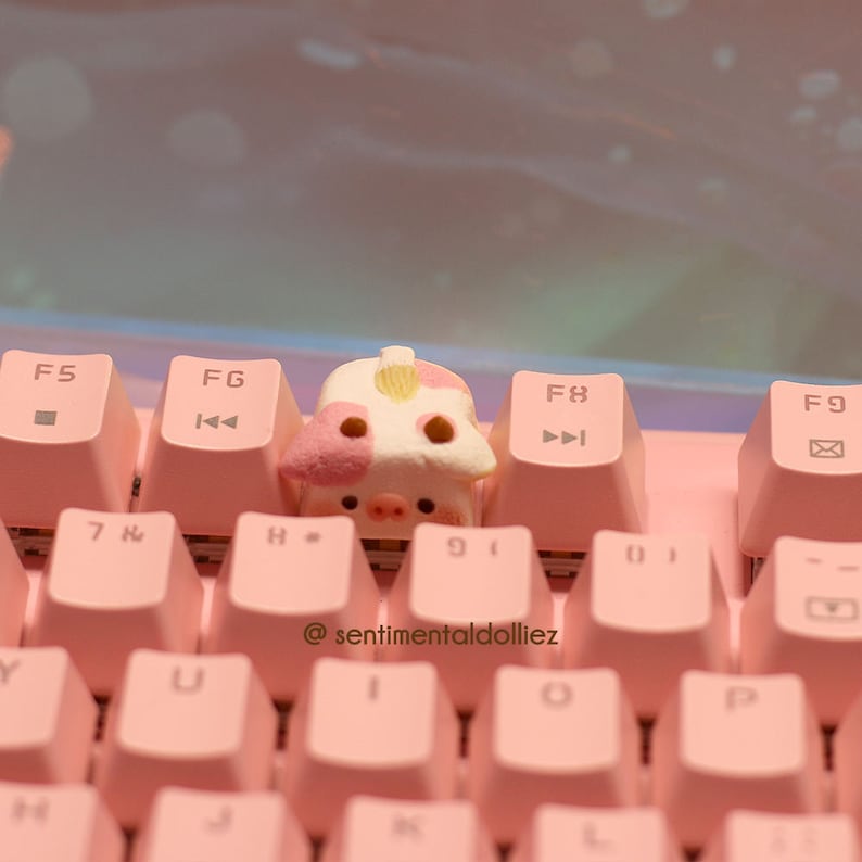 May include: A pink keyboard with a pig-shaped keycap. The keycap is white with pink spots and has a yellow nose. The keyboard has the keys F5, FG, F8, and F9 visible. The text "@sentimentaldolliez" is visible on the keyboard.