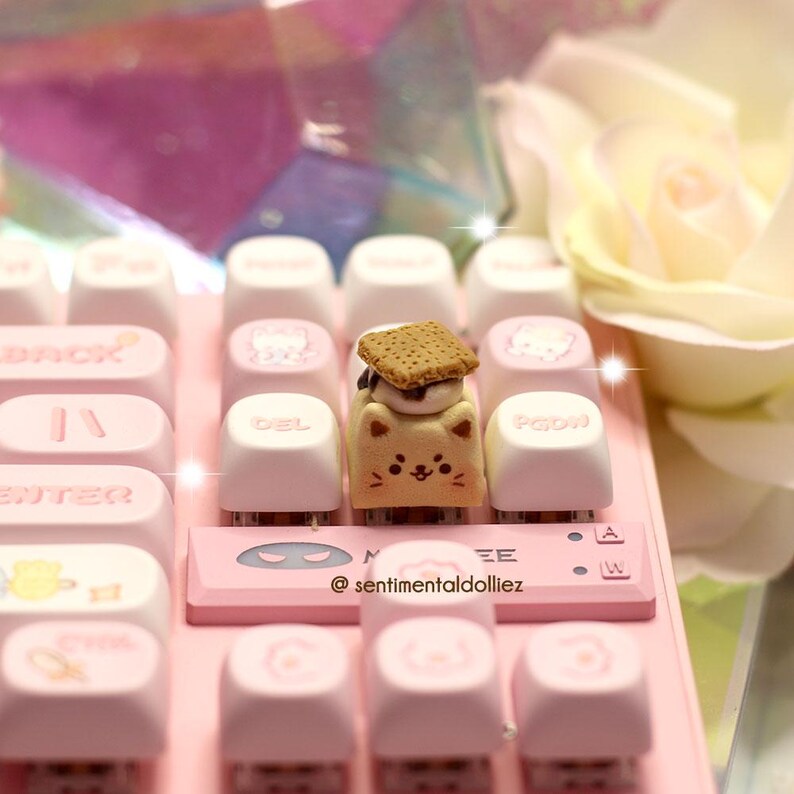 K&ouml;nnte beinhalten: Eine pinkfarbene mechanische Tastatur mit wei&szlig;en und pinkfarbenen Tastenkappen. Eine individuelle Tastenkappe in Form eines Katzen-S'more ist auf der Tastatur zentriert. Die Tastatur hat den Text "@ sentimentaldolliez" unten.