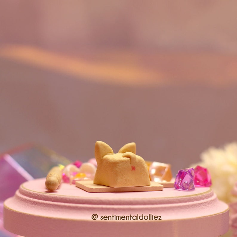 K&ouml;nnte beinhalten: Eine kleine Tonskulptur einer Katze mit rosa Ohren, die auf einer rosa Oberfl&auml;che mit einem Nudelholz und bunten Edelsteinen liegt. Die Oberfl&auml;che hat einen goldenen Rand und der Text "@sentimentaldolliez" ist darauf geschrieben.
