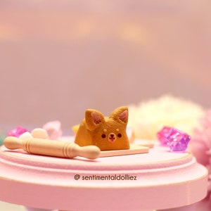 K&ouml;nnte beinhalten: Ein kleiner brauner und wei&szlig;er Kuchen in Hundeform auf einer rosa Oberfl&auml;che mit einem Nudelholz und violetten Kristallen. Der Kuchen hat ein s&uuml;&szlig;es Gesicht und liegt auf einem kleinen Holzbrett. Der Text "@sentimentaldolliez" befindet sich auf der Oberfl&auml;che.